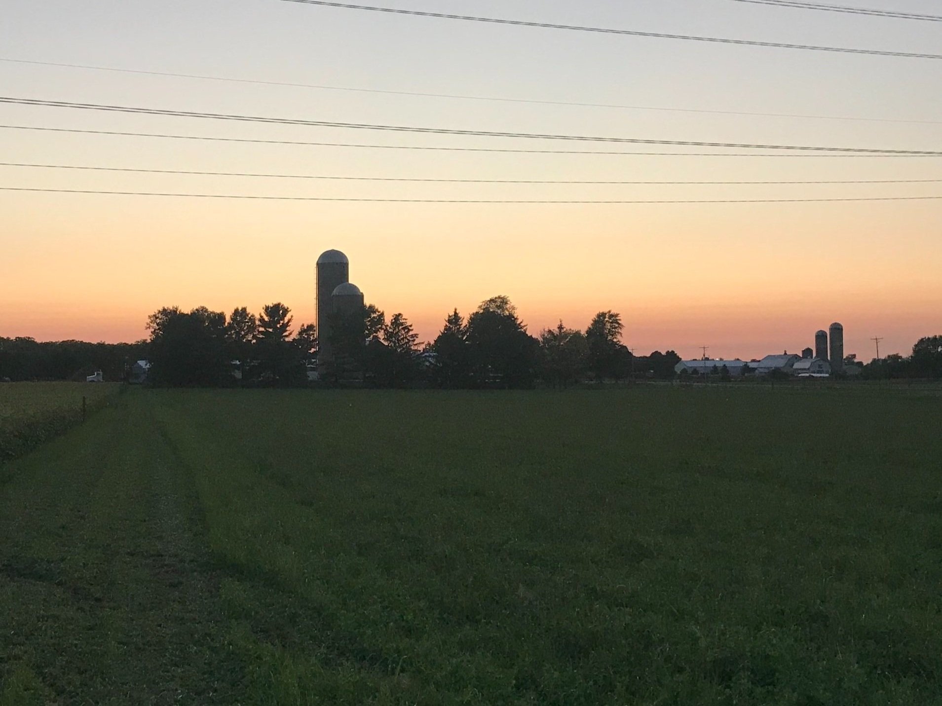 Sunset over a rural landscape featuring a green field, silos, and farm buildings in the distance, with power lines silhouetted against the colorful sky.