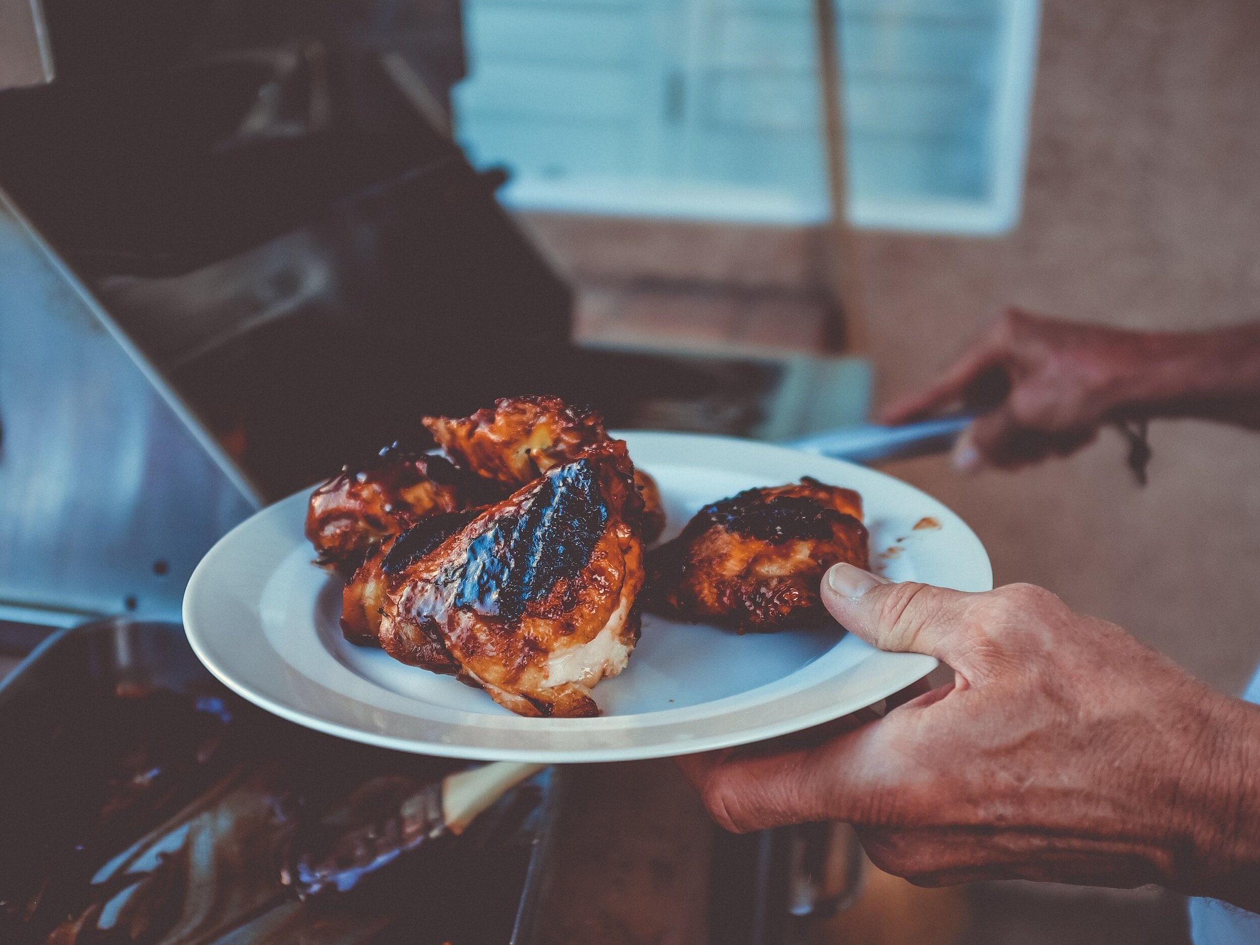 Grilled chicken thighs on a white plate held by a person, showcasing a delicious barbecue meal cooked on an outdoor grill.